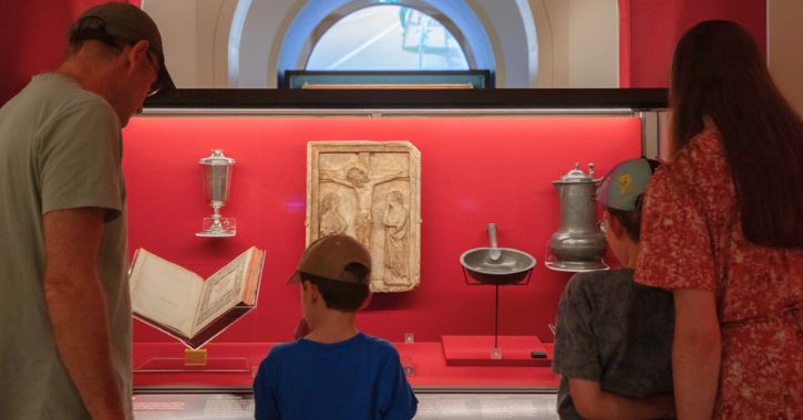 Family - two adults and 2 children stood in front of cabinet displaying historic Faith related objects inside the Faith Museum, Bishop Auckland.